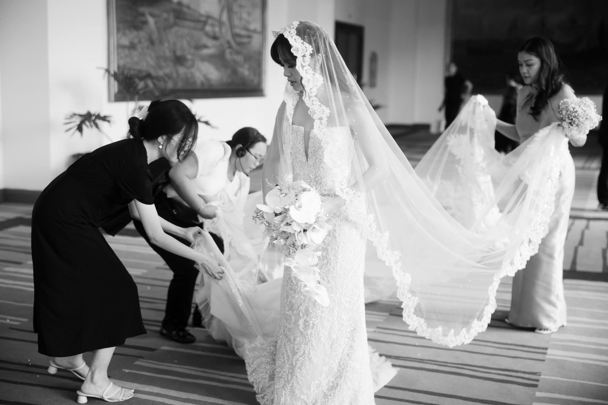 A bride in an intricately embroidered lace gown and cathedral-length veil, surrounded by her bridal team as they carefully arrange her train before the ceremony. Captured in timeless black and white, the moment celebrates the beauty of preparation and the artistry of bridal couture.