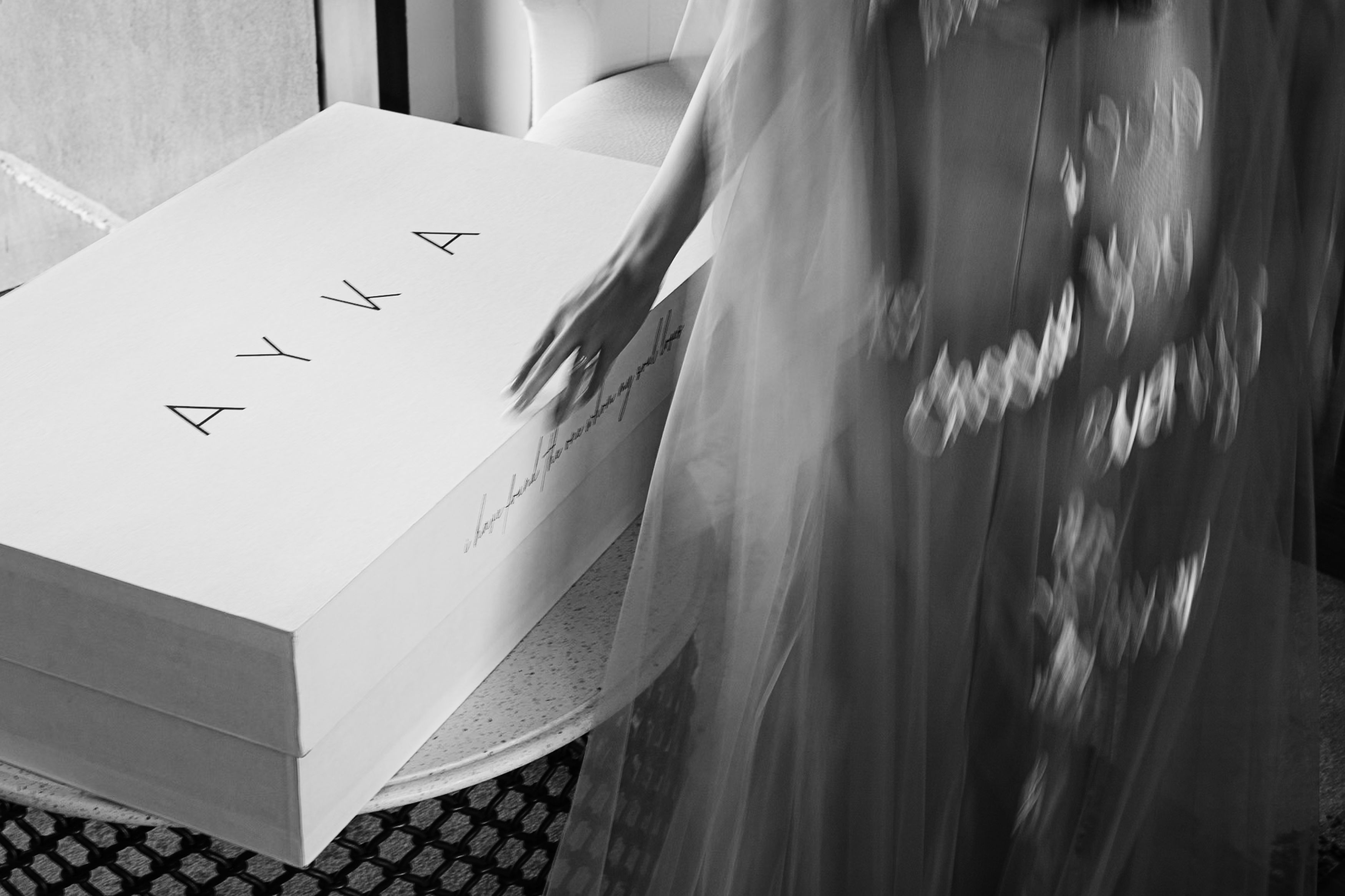 A black-and-white editorial photograph of a bride in an embroidered sheer veil, reaching toward a minimalist white box with elegant lettering, captured with soft motion blur for a timeless, artistic feel.