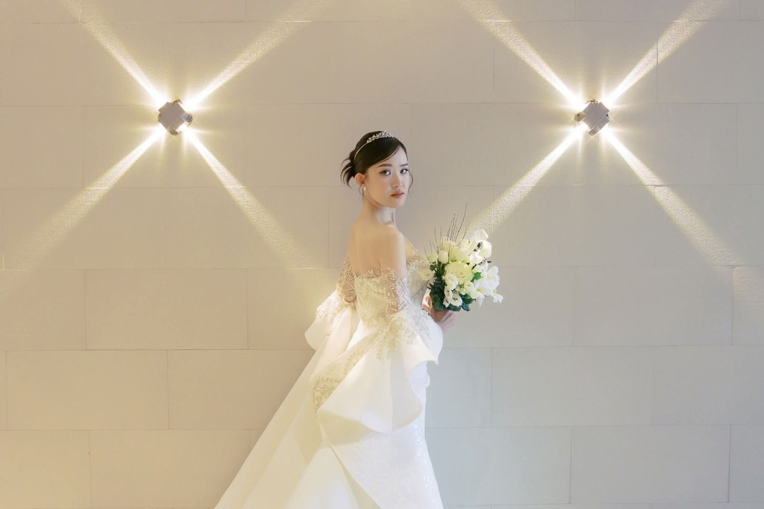 A bride in an off-shoulder ivory gown with beaded lace sleeves and a sculpted satin peplum, holding a white bouquet against a wall lit with star-shaped beams of light.