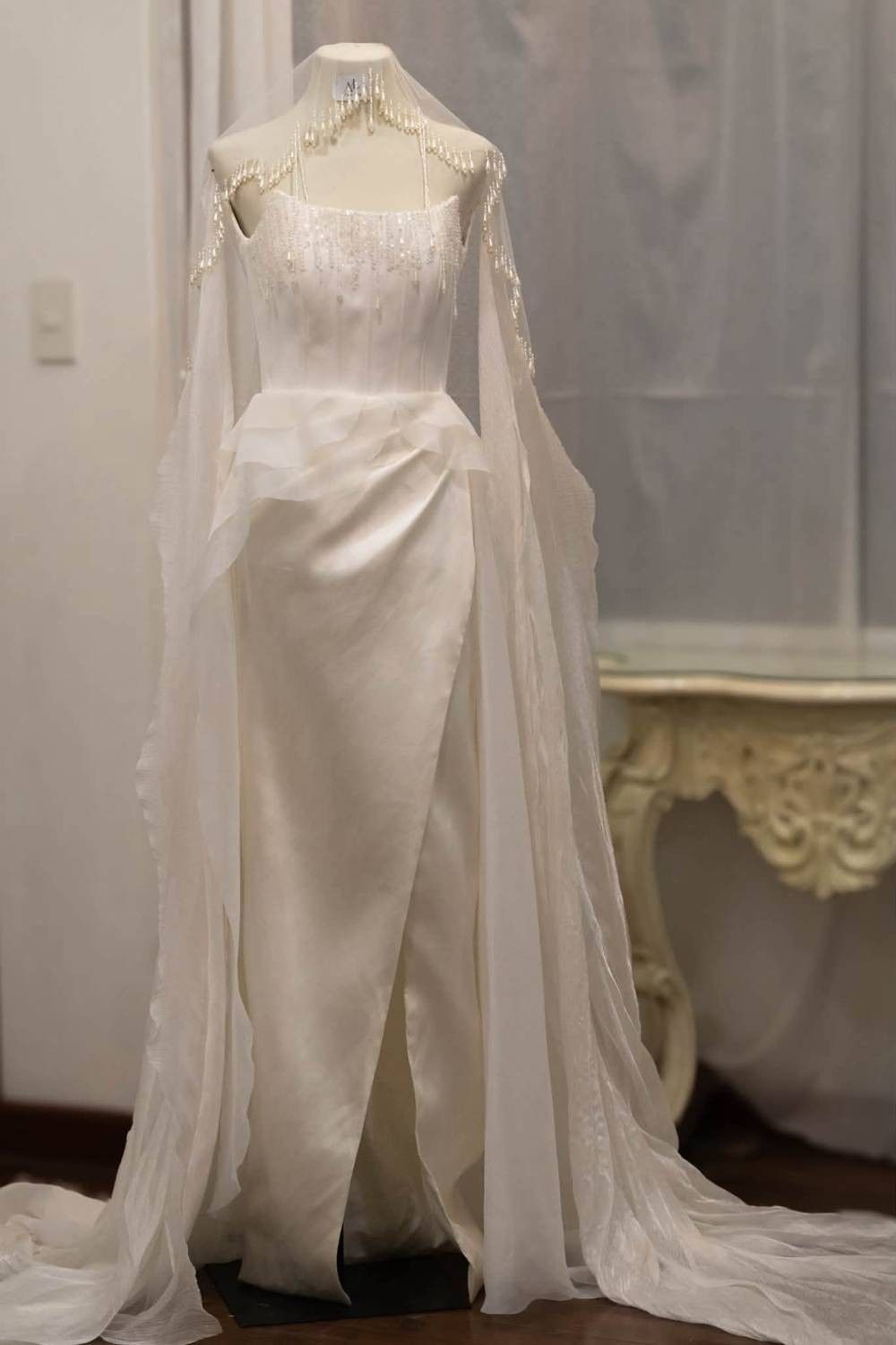 A bride walks down an outdoor garden aisle wearing an elegant ivory satin gown with a draped bodice, thigh-high slit, and delicate beaded mantilla veil, complemented by a flowing train and warm golden evening lights.