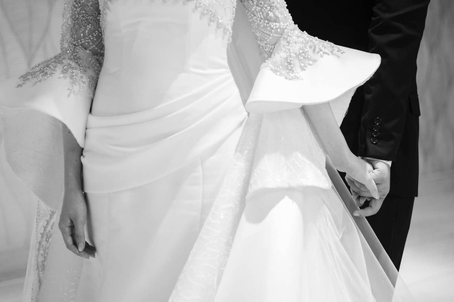 Black-and-white detail of an ivory bridal gown: sculpted satin sleeves edged with pearl beading, the bride's hand holding the groom's.